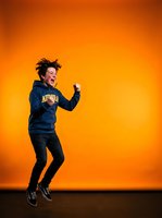 Excited young male with dark hair, Caucasian ethnicity, jumping with fist raised against vibrant orange gradient background, wearing navy hoodie and black pants, expressing joy and celebration for lifestyle and motivational content.