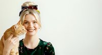 Young blonde woman of Caucasian ethnicity wearing green sequined top and burgundy sunglasses on head, making peace sign gesture with red nail polish, smiling at camera against white background.