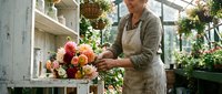 Mature woman florist arranging fresh orange and pink roses in greenhouse workspace, creating floral arrangements for events and celebrations.