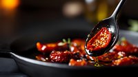 Roasted cherry tomatoes drizzled with olive oil in black cast iron pan, garnished with fresh herbs. Golden oil drips from spoon in moody lighting setting.