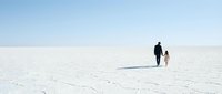 Adult male and young child walking together across vast white salt flat landscape under clear sky, depicting family bonding and exploration.