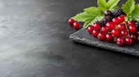 Fresh red currants and blackberries with green leaves arranged on black slate board against dark textured background with copy space for menu design.