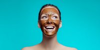 Young woman with chocolate facial mask laughing against turquoise background, showcasing skincare routine and joyful self-care moment.