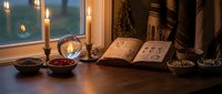 Candlelit tarot reading setup with open divination book, crystal ball, dried herbs and berries in bowls on wooden table at dusk for spiritual practices.