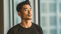 Asian man with eyes closed in black t-shirt standing by window, bathed in warm natural light, expressing peaceful contemplation in modern interior setting.