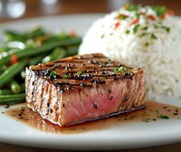 Juicy grilled beef steak with grill marks served medium rare alongside steamed white rice and fresh green beans, garnished with herbs and sauce on white plate.