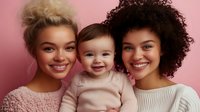 Happy diverse family portrait with smiling mothers and baby against pink background. Multiracial women with blonde and curly hair holding infant child.