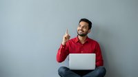 Young Indian businessman in red shirt pointing up with laptop, sitting against gray wall background, having creative idea or eureka moment.
