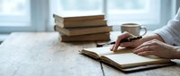 Woman writing in notebook at desk with stack of books and coffee cup in bright natural light creating peaceful study atmosphere for education and learning.