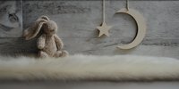 Knitted bunny toy sitting on soft white fur with hanging wooden moon and star decorations against rustic gray wall. Cozy nursery decor concept.