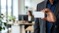 Businessman of Middle Eastern descent in dark blazer and blue shirt holding blank white business card in modern office environment with colleagues and plants in background.