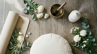 Flat lay overhead view of wellness and spa essentials with white flowers, green foliage, mortar and pestle, candle, and rolled paper on wooden surface.