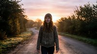Young woman with long brown hair wearing a dark jacket and beanie standing on a rural dirt road at golden hour sunset, surrounded by trees and natural landscape.