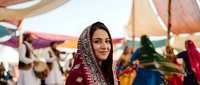 Young South Asian woman in traditional burgundy embroidered sari and headscarf smiling at colorful outdoor market festival with fabric canopies and vendors in background.