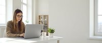 Young professional woman with glasses working on laptop at modern white desk in bright office space with natural light and minimalist decor.