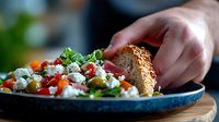 Fresh Mediterranean salad with feta cheese, tomatoes, and olives served on blue ceramic plate with sesame bread, close-up view showing vibrant colors and textures.