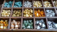 Colorful mineral crystals and gemstones displayed in vintage wooden compartment box, arranged by color including turquoise, gold, amber and silver specimens.