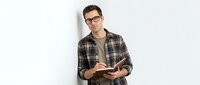 Young man with glasses wearing plaid shirt holding open book against white wall, suitable for education, learning, and literacy concepts.