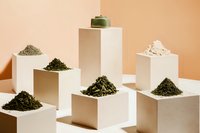 Loose tea leaves and powder displayed on minimal white geometric podiums against peachy background, creating modern composition for tea packaging and marketing.