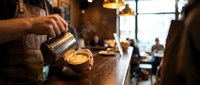 Skilled barista pouring steamed milk into a latte art cup at a warm, rustic coffee shop counter with soft ambient lighting.