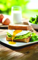 Fresh breakfast sandwich with poached egg, avocado slices, and lettuce on toasted bread, served on white plate with glass of milk and eggs in background.