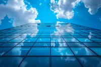 Modern glass office building facade reflecting bright blue sky and white clouds, photographed from ground level looking upward. Architectural perspective.