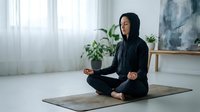 Young Caucasian woman in black hoodie meditating in lotus pose on yoga mat in bright minimalist home interior with plants.