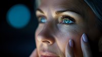 Young Caucasian woman with bright blue eyes in dramatic dark lighting touches her face gently, showing emotion and vulnerability in atmospheric night portrait.