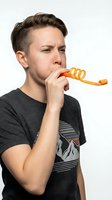 Young male with light skin tone and dark hair wearing casual dark t-shirt blowing orange party horn against white background for celebration and festive event concepts.