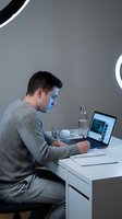 Young Caucasian businessman in gray sweater working on laptop at modern white desk with ring light in contemporary office workspace for remote work.