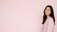 Young woman with long black hair and South Asian ethnicity, wearing a light pink button-up shirt, smiling at camera against soft pink background. Suitable for lifestyle, wellness, and professional content.
