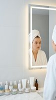 Young woman in white bathrobe and towel examining face in illuminated bathroom mirror with skincare products on marble counter for morning routine.