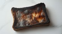 Burnt toast slice with charred black edges and golden brown center on white marble surface for food photography and culinary concepts.