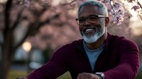 Mature African American man with gray beard wearing burgundy sweater and glasses smiling outdoors during spring sunset with cherry blossoms in background.