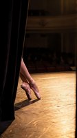 Ballet dancer legs in pointe shoes on wooden stage floor with dramatic lighting and black curtain backdrop for performing arts and dance concepts.