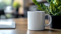 White ceramic coffee mug on wooden desk with green plant and notebook in background, modern minimalist workspace setting with soft natural lighting.