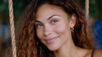 Young mixed race woman with curly brown hair and natural makeup smiling warmly outdoors near rope swing, expressing genuine happiness and confidence in casual summer setting.