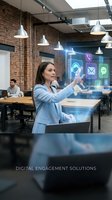 Woman in light blue blazer interacting with holographic digital interface displaying communication and app icons in modern brick office workspace with colleagues and pendant lighting.