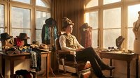 Young male fashion designer working on laptop in sunlit atelier surrounded by vintage clothing and accessories for creative inspiration and design process.