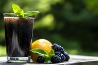 Fresh blackberry smoothie in glass with mint garnish, lemon and raw berries on wooden surface against blurred green background.