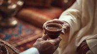Person in traditional Middle Eastern attire holding small cup of coffee or tea against backdrop of ornate carpet and wooden furniture.