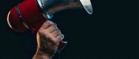 Male hand holding red metallic megaphone against dark background for announcements, marketing campaigns, and promotional communications in business.