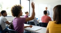 African American female student raising hand in diverse classroom with teacher and classmates during interactive learning session.