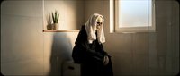 Mature woman with long blonde hair wearing black clothing sits pensively by window in minimalist bathroom interior with warm lighting and potted plant.