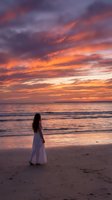 Woman with long dark hair wearing white flowing dress standing on sandy beach gazing at dramatic orange and purple sunset sky over ocean waves. Perfect for travel, lifestyle, and contemplation themes.