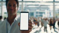 Caucasian businessman showing blank smartphone screen in busy airport terminal with blurred crowd in background, mockup for app or mobile content.