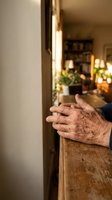Elderly hands resting on wooden kitchen counter in warm cozy home interior with soft lighting and blurred background for lifestyle content.