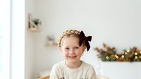 Young girl with reddish-blonde hair wearing cream sweater and brown bow, smiling at camera in bright room with holiday decorations and garland in background.
