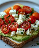 Fresh avocado toast topped with cherry tomatoes, crumbled feta cheese and herbs on whole grain bread, garnished with black pepper and seasonings.