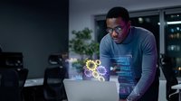 Young African American man working with holographic gear interface on laptop in modern office environment showcasing futuristic technology and innovation.